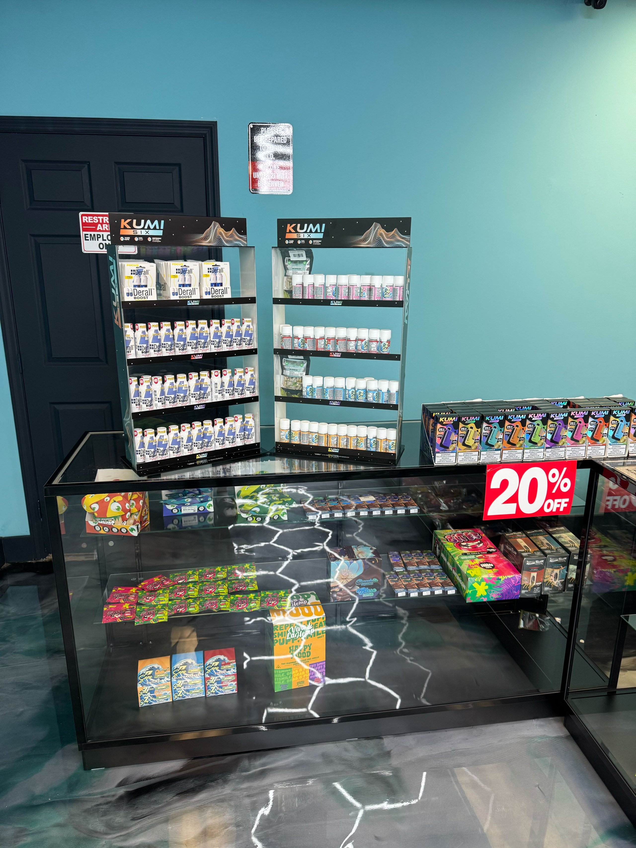 Vape shop display with KUMI branded shelving units containing various e-liquid bottles and vaping products against turquoise wall.