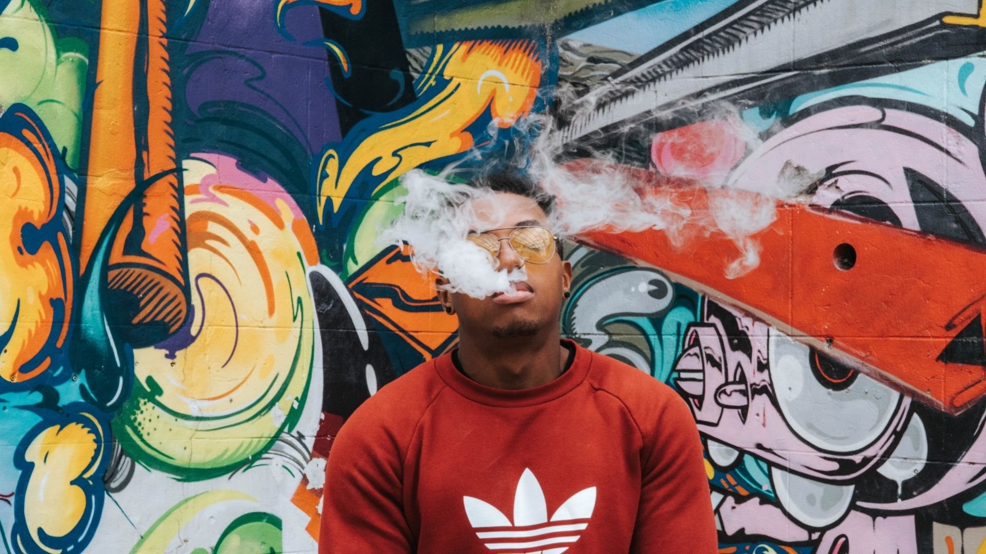 Person in red shirt exhaling smoke in front of colorful graffiti mural