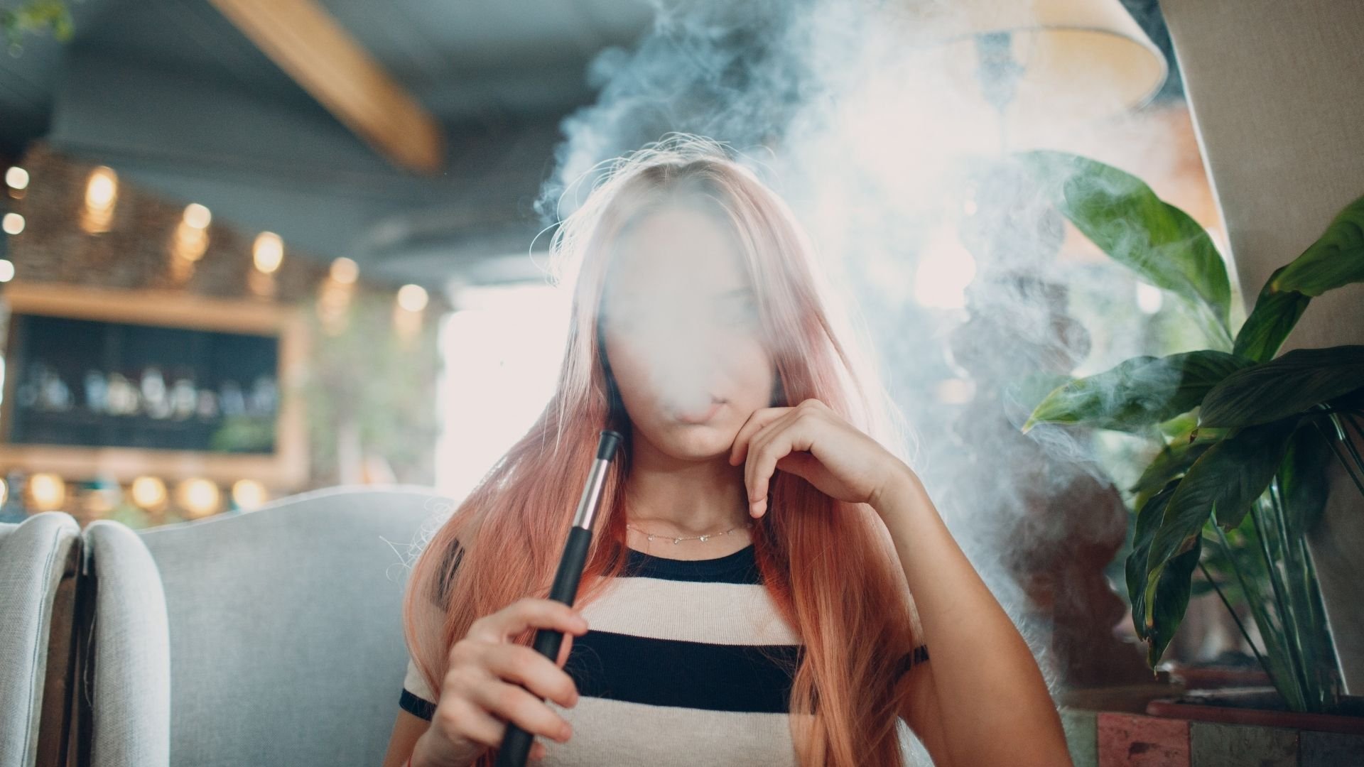 Woman with reddish-brown hair vaping indoors surrounded by plants and warm lighting.